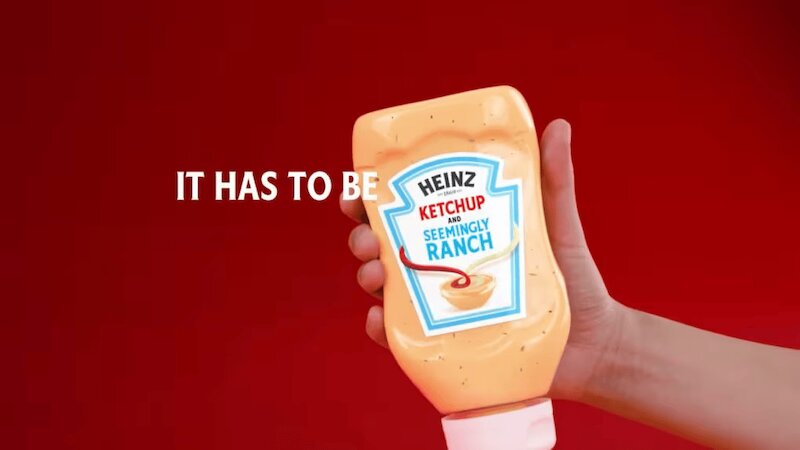 A hand holding a bottle of Heinz Ketchup and Seemingly Ranch sauce against a solid red background.